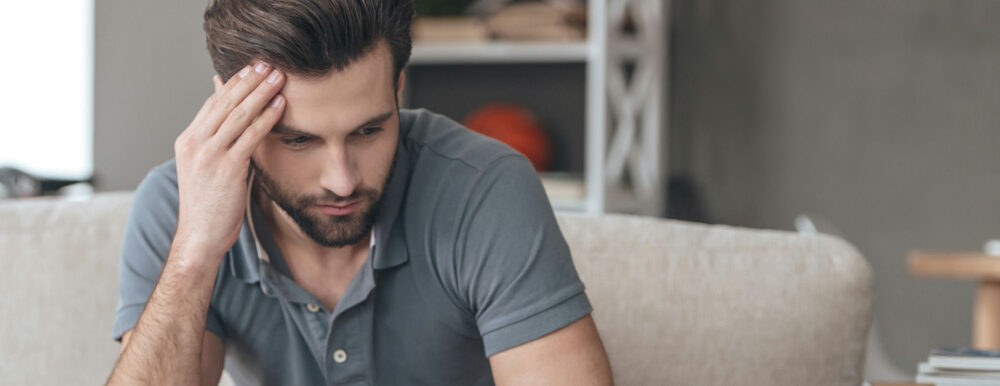 man sitting on a couch with his hand on his head, having difficulty thinking through brain fog